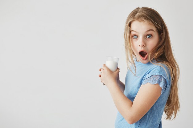 Close up portrait of funny good-looking little girl with light hair  with shocked expression, holding glass of milk with hands.