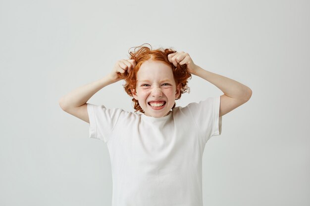 Close up portrait of funny little boy with ginger hair and freckles smiling with teeth, having happy expression, holding hair with hands.