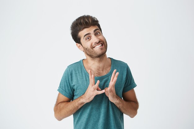 Close up portrait of funny stylish spanish guy in blue tshirt, biting lips and gesticulating with hands expressing feeling super awkward. Body language.