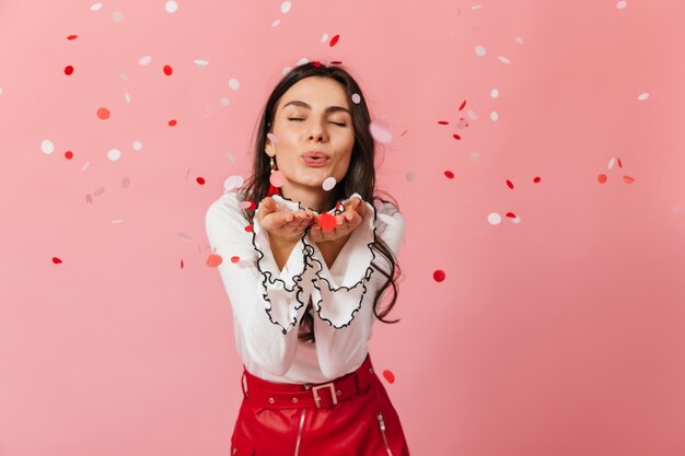 Close-up portrait of girl in leather skirt sending air kiss on pink background with confetti.