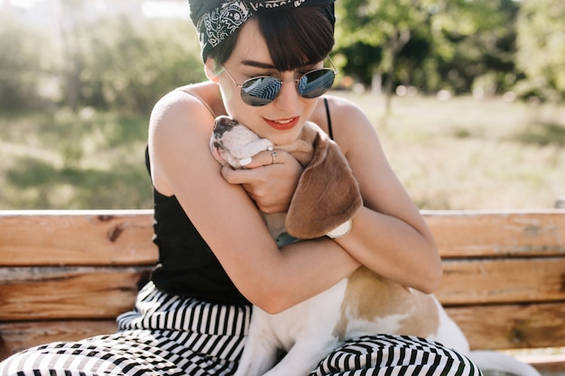 Портрет крупным планом радостной брюнетки, обнимающей ее гончую собаку с нежной улыбкой.