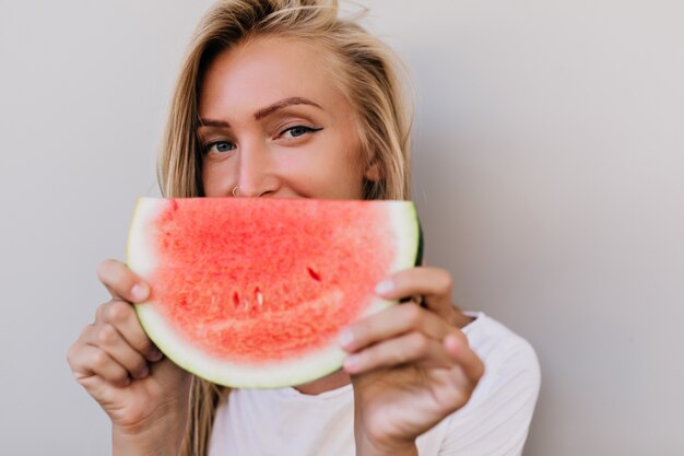 Close-up portrait of glad caucasian woman eating fruits. Indoor photo of adorable blonde woman fooling around on light background.