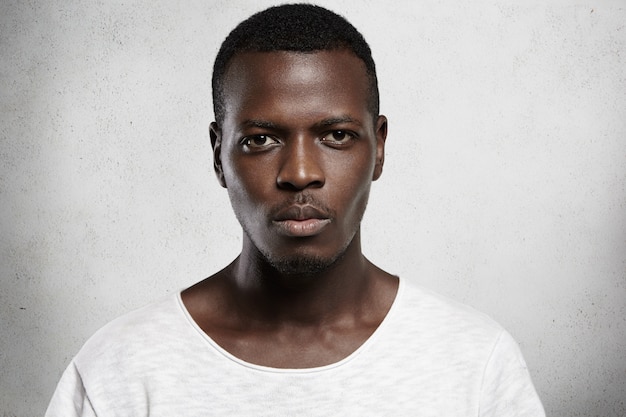 Close up portrait of good-looking serious African man with healthy clean skin wearing white casual t-shirt posing isolated against gray  wall with copy space for your promotional content