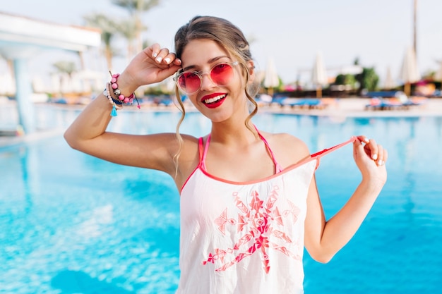 Close-up portrait of good-looking tanned girl in pink sunglasses posing with hands up and happy face expression