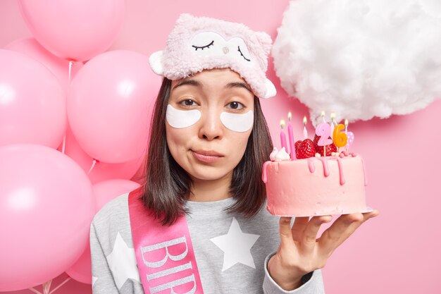 Close up portrait of good looking young Asian female model applies patches under eyes wears sleepmask and pajama celebrates birthday