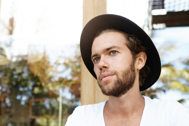 Close up portrait of good-looking young Caucasian hipster with thick beard wearing black hat and white t-shirt looking sad or tired