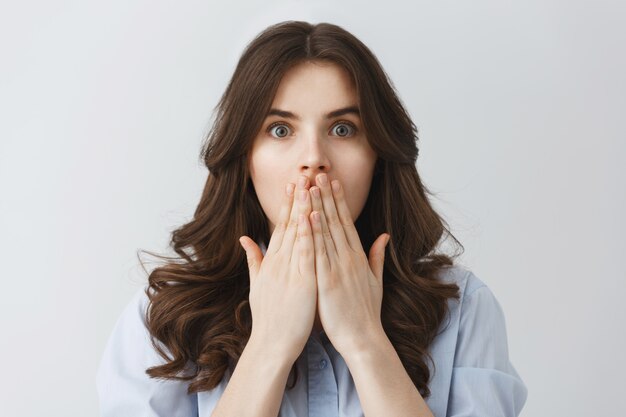 Close up portrait of good-looking young student girl with dark wavy hair closing mouth with hands in frightened look watching scary movie with her boyfriend.