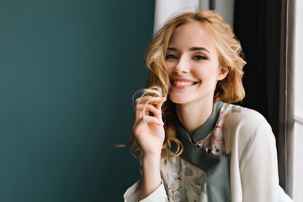 Close-up portrait of gorgeous blonde young woman wearing turquoise pajamas, sitting next to the window. She's happy, smiling, touching her wavy hair.