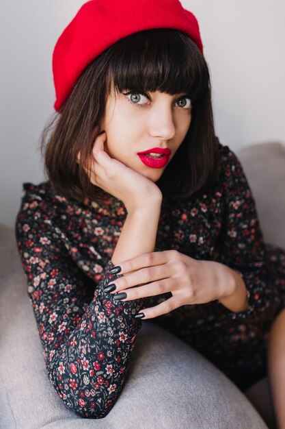 Close-up portrait of gorgeous french girl with short haircut in red beret posing with surprised face expression. Adorable  young woman in vintage attire sits in a chair, propping her cheek with hand.