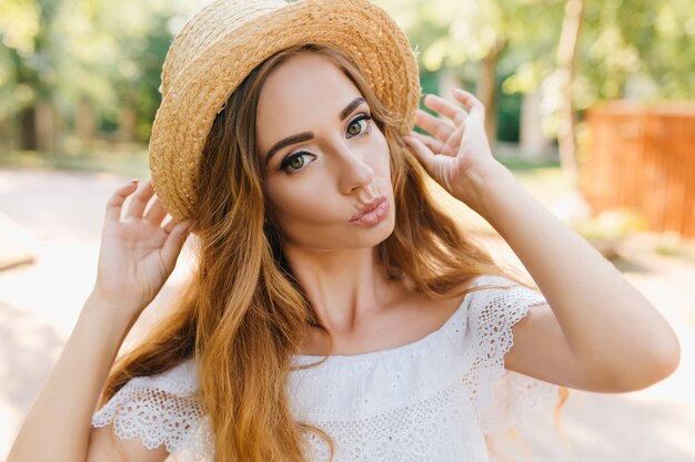 Close-up portrait of gorgeous long-haired girl with green eyes posing with pleasure in sunny day. Photo of fair-haired lady with kissing face expression wearing ring.