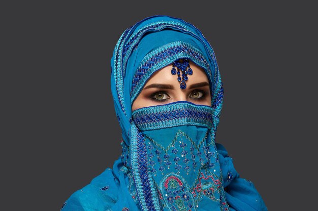 Close-up portrait of a gorgeous young woman with beautiful smoky eyes wearing an elegant blue hijab decorated with sequins and jewelry. She is posing sideways and looking at the camera on a dark backg