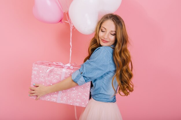 Close-up portrait of graceful curly girl with lovely face holding present and balloons for friend's birthday. Charming long-haired young woman with eyes closed in stylish attire received gift at party