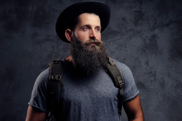 Close-up portrait of a handsome bearded traveler in hat with backpack. Isolated on a dark background.