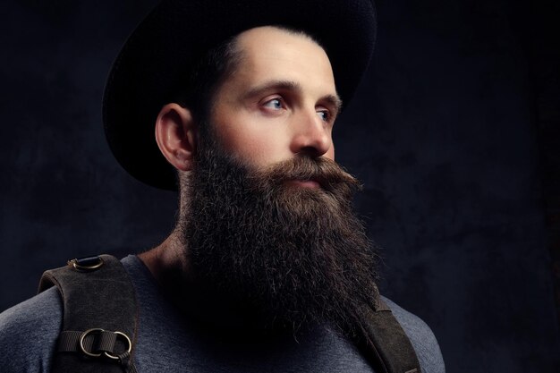Close-up portrait of a handsome bearded traveler in hat with backpack. Isolated on a dark background.