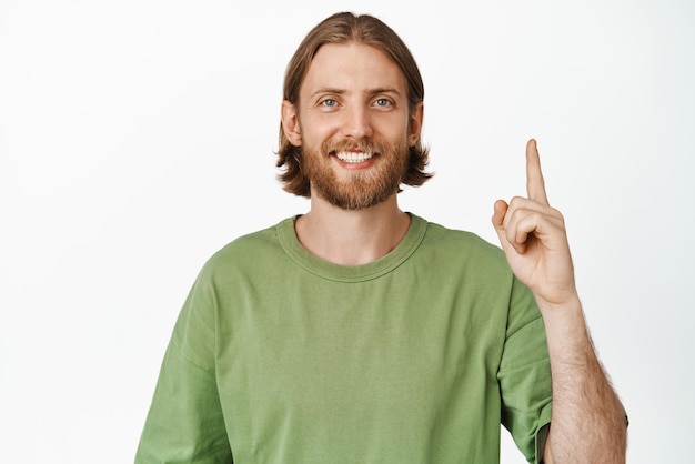 Close up portrait of handsome blond man in tshirt pointing finger up and smiling pleased showing advertisement logo banner standing against white background