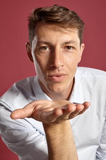 Close-up portrait of a handsome brunet man with brown eyes, wearing in a white shirt and a terracotta tie. He is sending a kiss while posing in a studio against a red background. Concept of gesticulat