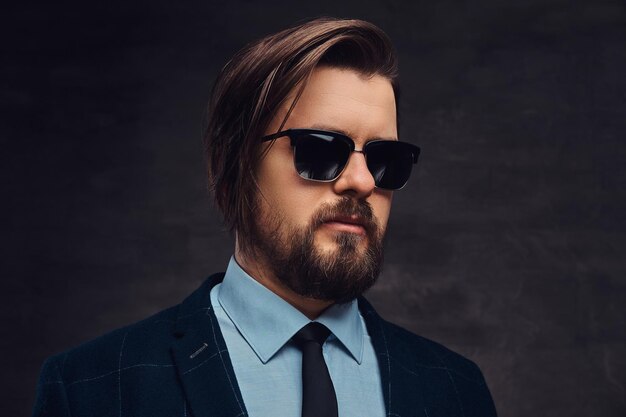 Close-up portrait of a handsome fashionable middle-aged man with beard and hairstyle dressed in an elegant formal suit and sunglasses on a textured dark background in studio.