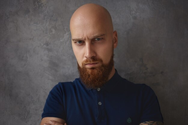 Close up portrait of handsome frowning young bald man with thick beard having grumpy sullen look with eyes full of anger and rage. Negative human facial expressions and emotions