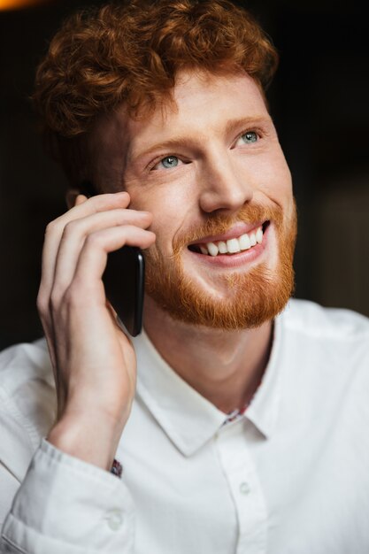 Close-up portrait of handsome smiling redhead bearded man in white shirt tolking on mobile phone