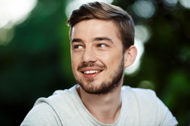 Close up portrait of handsome smiling young man in white t-shirt looking away on blurry outdoor nature