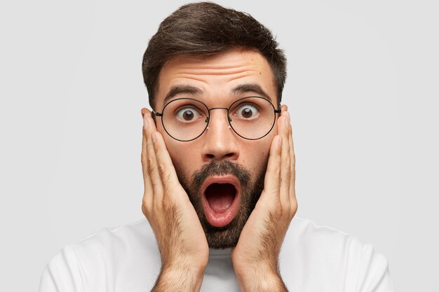 close up portrait of handsome terrified young male with stubble being shocked to bottom of his heart, touches cheeks with both palms, notices something unexpected, isolated over white wall