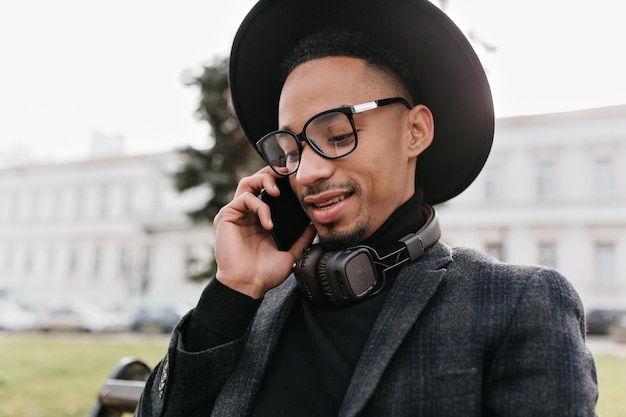 Крупным планом портрет красивый молодой африканский человек разговаривает по телефону. Открытый выстрел темнокожего парня в модной шляпе, звонящего кому-то.