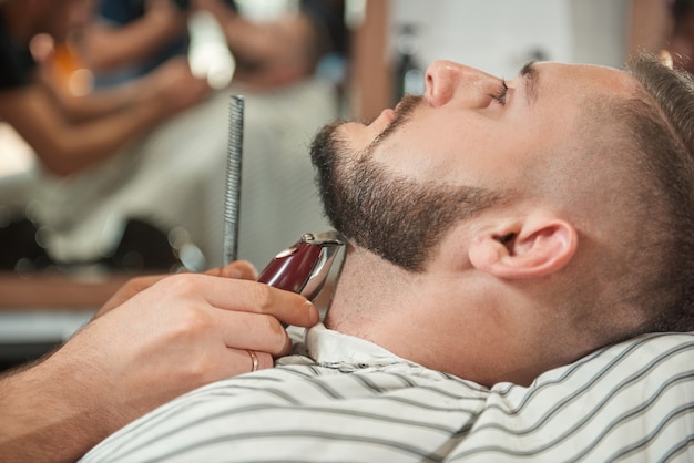 Chiuda sul ritratto di un bel giovane barbuto che ottiene la sua barba rifilata da un barbiere professionista.