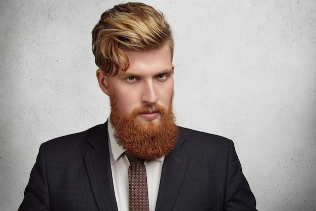 Free photo close up portrait of handsome young corporate worker with stylish haircut and thick beard wearing elegant suit looking  with serious and focused face expression before business meeting