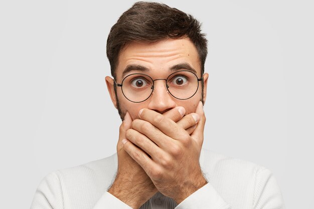 Close up portrait of handsome young man covers mouth with both palms, tries to be mute, stops shouting