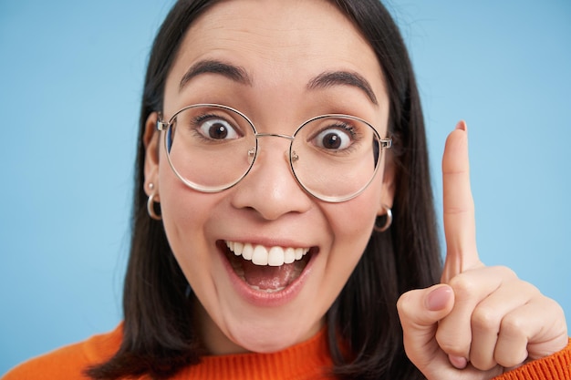 Free photo close up portrait of happy asian girl in glasses raises finger has revelation suggest great idea say