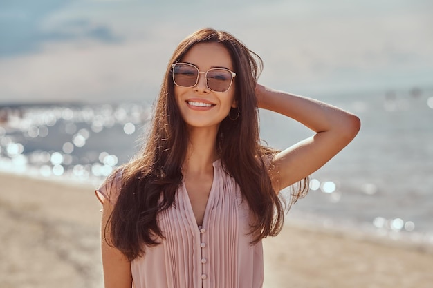 해변에서 선글라스와 드레스에 긴 머리를 가진 행복 한 아름 다운 갈색 머리 소녀의 클로 우즈 업 초상화.