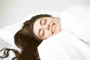 Free photo close up portrait of happy carefree young caucasian woman with shiny brunette hair closing eyes with pleasure, relaxing in bed wrapped in white blanket, feeling warm and comfortable on cold winter day