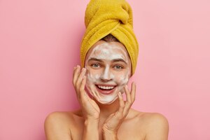 Close up portrait of happy caucasian woman washes face with facial soap and water, wants to have healthy complexion, removes dirt and sweat sebum, yellow wrapped towel on head