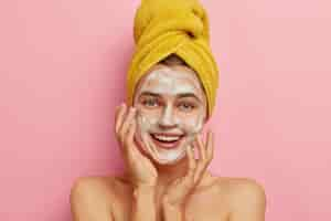 Free photo close up portrait of happy caucasian woman washes face with facial soap and water, wants to have healthy complexion, removes dirt and sweat sebum, yellow wrapped towel on head