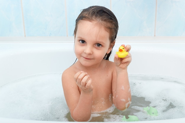 Close up portrait of happy charming little girl sitting in bath tub plays with yellow duck in bathroom.
