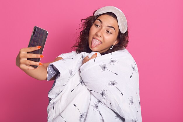 Close up portrait of happy female wrapped white blanket with dandelion, showing her tongue, making selfie