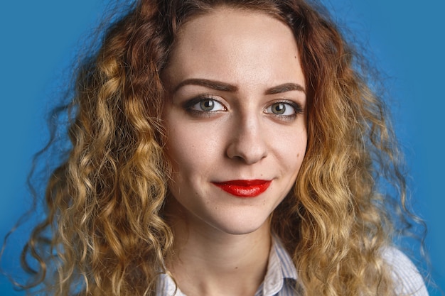 Close up portrait of happy friendly looking young lady with voluminous wavy hair and red lips relaxing indoors against blue wall wall, having joyful positive expression on her pretty face