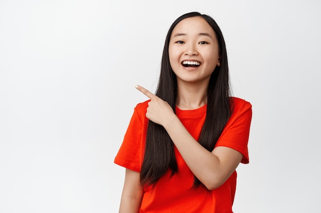 Крупным планом портрет счастливой корейской девушки, указывающей пальцем в верхнем левом углу со счастливой улыбкой на белом