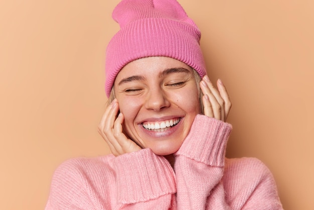 행복한 사랑스러운 여성의 클로즈업 초상화는 얼굴에 손을 대고 즐겁게 눈을 감고 있는 모습을 보여주며 하얀 치아가 좋은 분위기에 분홍색 모자와 스웨터를 입고 베이지색 스튜디오 벽에 격리되어 있습니다.