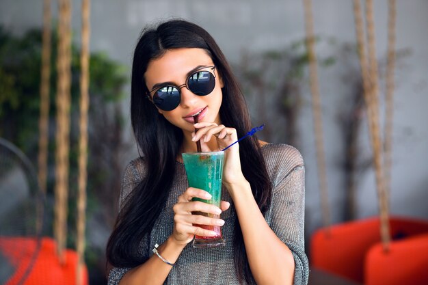 Close up portrait of happy pretty brunette woman drinking tasty cold cocktail, stylish outfit and mirrored sunglasses, enjoy her weekend, party time .