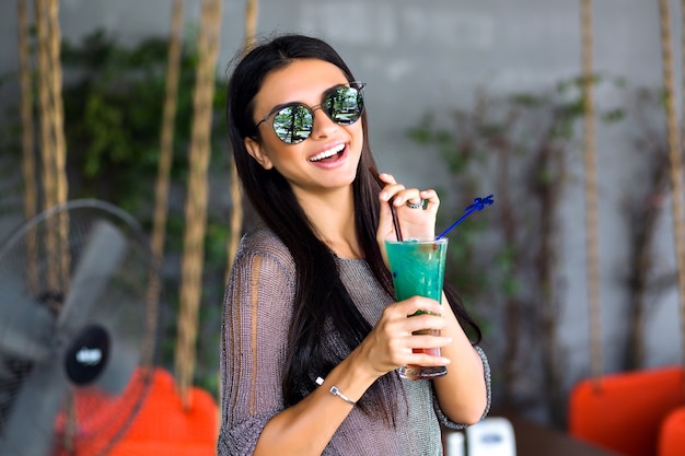 Close up portrait of happy pretty brunette woman drinking tasty cold cocktail, stylish outfit and mirrored sunglasses, enjoy her weekend, party time .