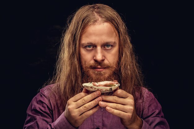 Close-up portrait of a happy redhead hipster male with long hair and full beard dressed in a purple shirt holds a small saucer with tea, looking at a camera. Isolated on a dark textured background.