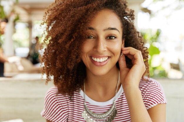 Close up portrait of happy smiling woman with broad smile has delighted expression, rests against cafe interior alone, being in good mood, has break after hard working day. Positive emotions concept