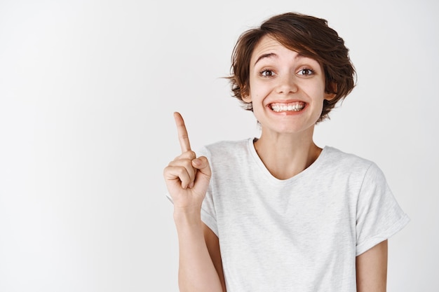 Close up portrait of happy smiling woman without makeup, wearing t-shirt and pointing up, showing, standing against white wall