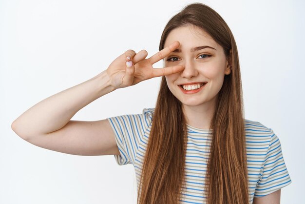 Close up portrait of happy young woman smiling with white teeth showing peace vsign wearing striped tshirt white background