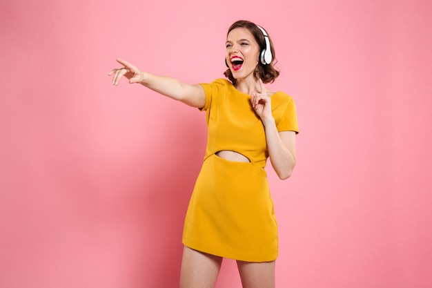 Close-up portrait of happy young woman in yellow dress and headphones pointing with finger, looking aside