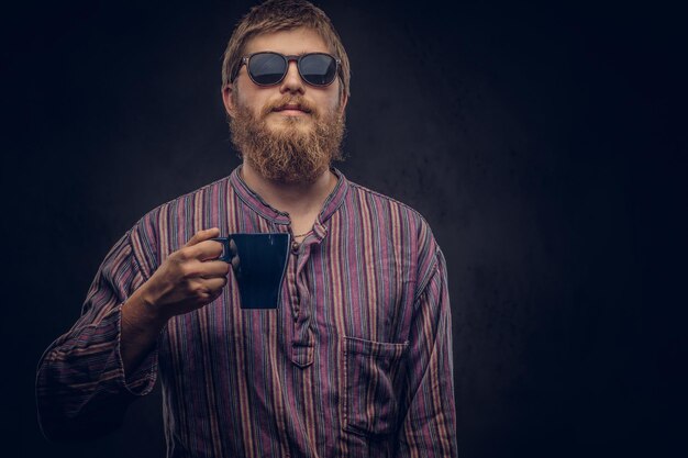 昔ながらのシャツを着たサングラスをかけている流行に敏感な男のクローズアップの肖像画は、コーヒーのカップを保持しています。暗い背景に分離されています。