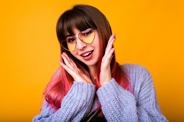 close up portrait of hipster pretty woman, surprised exited emotions, hearted glasses and cozy sweater, unusual ombre fuchsia hairs, yellow wall.