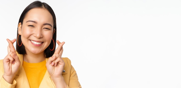 Close up portrait of hopeful asian girl wishing cross fingers for good luck praying and smiling standing over white background