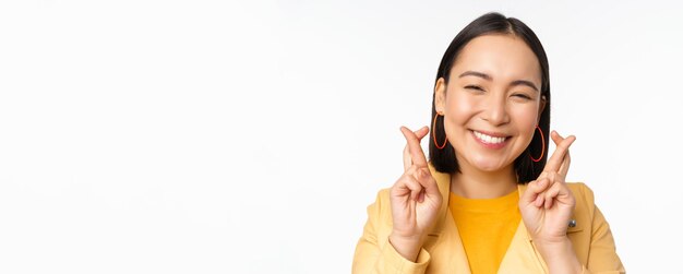 Крупным планом портрет обнадеживающей азиатской девушки, желающей скрестить пальцы на удачу, молящейся и улыбающейся, стоя на белом фоне